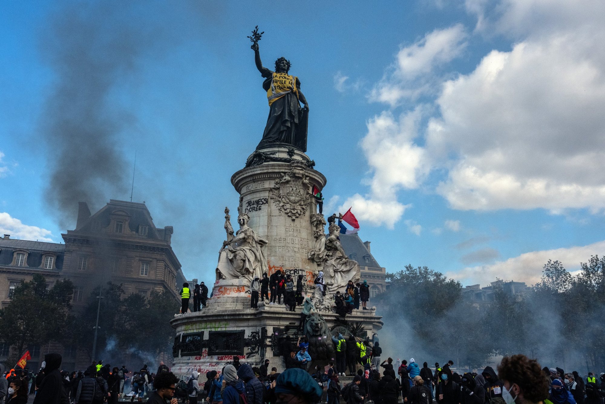 Διαδηλωτές πάνω και γύρω από το άγαλμα της Μαριάν στην Place de la République του Παρισιού, μέσα σε καπνούς, με σημαίες και συνθήματα στο βάθρο.